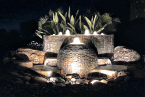 A brightly lit fountain surrounded by lush plants, illuminated against the night sky.