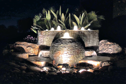 A brightly lit fountain surrounded by lush plants, illuminated against the night sky.