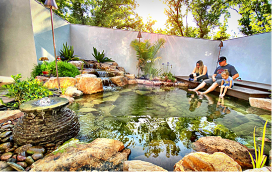 A family enjoys time together in a backyard pond, surrounded by a gentle waterfall and lush greenery.