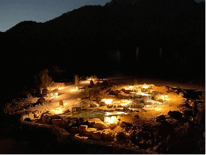 A serene night view of a hot spring, steam rising against a starry sky, surrounded by dark silhouettes of trees.