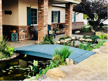 A small wooden bridge crosses over a backyard pond with water plants, next to a brick porch and a landscaped garden.