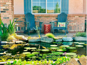 Two Adirondack chairs and a red side table sit on a patio behind a small pond with lily pads and aquatic plants in front of a brick house.